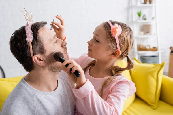 Girl in toy crown doing makeup with cosmetic brushes to father — Stock Photo