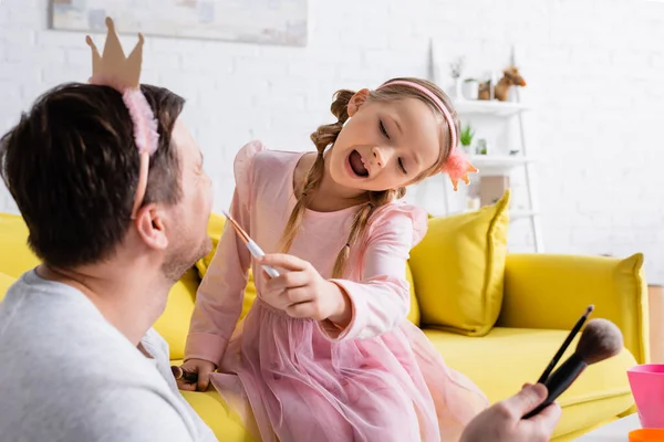 Homem em coroa de brinquedo segurando escovas cosméticas enquanto a filha excitada aplicando maquiagem, foreground borrado — Fotografia de Stock