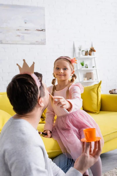 Excité fille faire maquillage à père tenant jouet tasse sur flou premier plan — Photo de stock