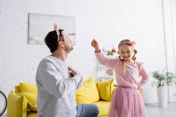 Man in toy crown holding hands on chest while inviting cheerful daughter to dance — Stock Photo