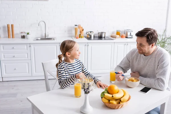 Homem alegre e filha olhando um para o outro durante o café da manhã na cozinha — Fotografia de Stock