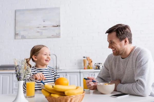 Padre e hija sonriendo el uno al otro mientras comen hojuelas de maíz cerca de frutas frescas en primer plano borroso — Stock Photo