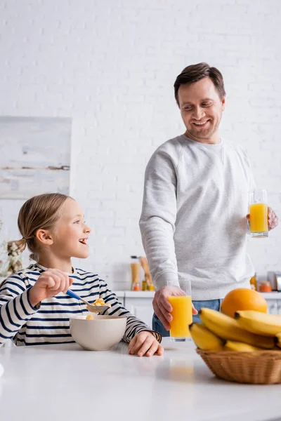 Sonriente hombre sosteniendo jugo de naranja cerca de hija comiendo copos de maíz para el desayuno — Stock Photo