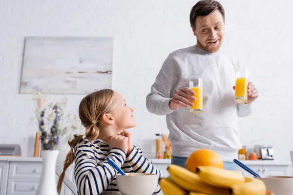 Pai alegre segurando suco de laranja perto da filha e frutas em primeiro plano desfocado — Fotografia de Stock