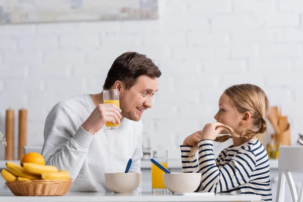 Улыбающийся мужчина держит стакан апельсинового сока во время разговора с дочерью во время завтрака — стоковое фото