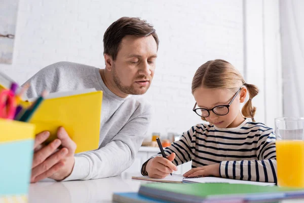 Père tenant un livre près de sa fille écrivant dans un cahier tout en faisant ses devoirs, au premier plan flou — Photo de stock