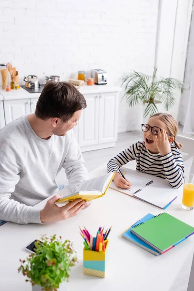 Man holding book near excited daughter doing homework in kitchen — Stock Photo