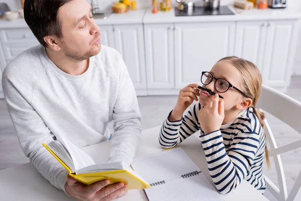 Menina brincalhão segurando caneta perto do nariz ao fazer lição de casa com o pai — Fotografia de Stock