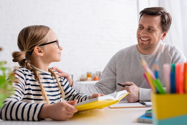 Sonriente hombre mirando a su hija sosteniendo el libro de texto mientras hace la tarea en primer plano borroso - foto de stock