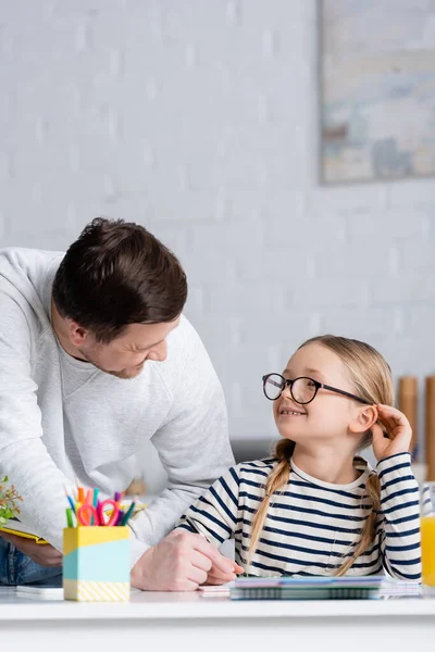 Sorrindo menina em óculos olhando para o pai ao fazer lição de casa, foreground borrado — Fotografia de Stock
