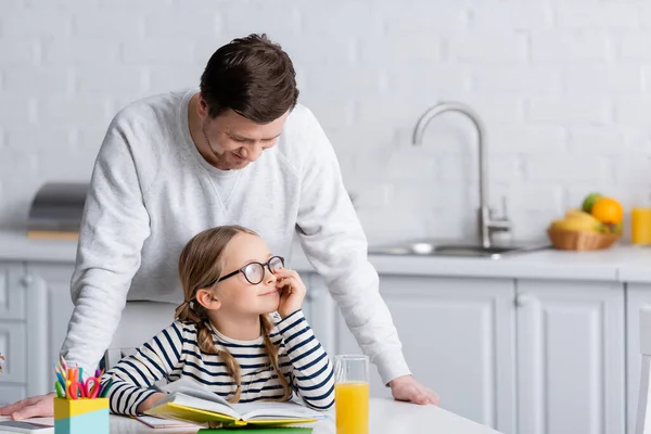 Чоловік стоїть біля усміхненої дочки робить домашнє завдання на кухні — стокове фото