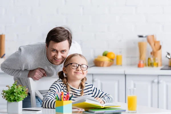 Heureux père regardant caméra près de sourire fille faire des devoirs dans la cuisine — Photo de stock
