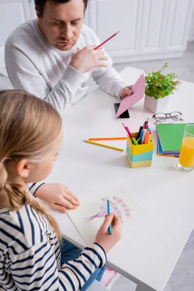 Child drawing happy mothers day card near father, blurred foreground — Stock Photo