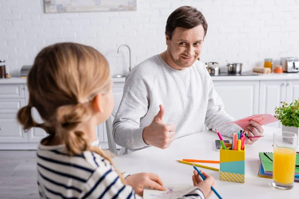 Homme joyeux montrant pouce vers le haut près de fille tenant crayon de couleur sur le premier plan flou — Photo de stock