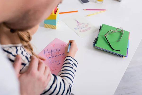 Partial view of dad touching shoulder of daughter drawing happy womens day card, blurred foreground — Stock Photo