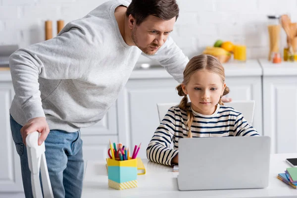 Chica haciendo la tarea cerca de portátil y padre en la cocina - foto de stock