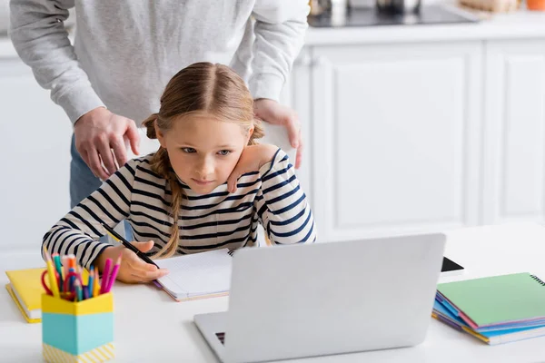 Father standing behind schoolgirl looking at laptop during online lesson, blurred foreground — Stock Photo