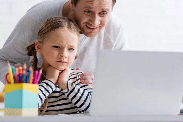 Lächelnder Mann berührt nachdenkliche Tochter während Online-Unterricht am Laptop auf verschwommenem Vordergrund — Stockfoto