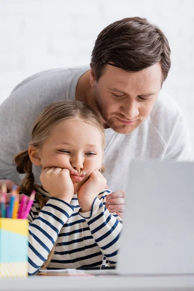 Тато торкається виснаженої дочки під час онлайн уроку вдома, розмитий передній план — стокове фото