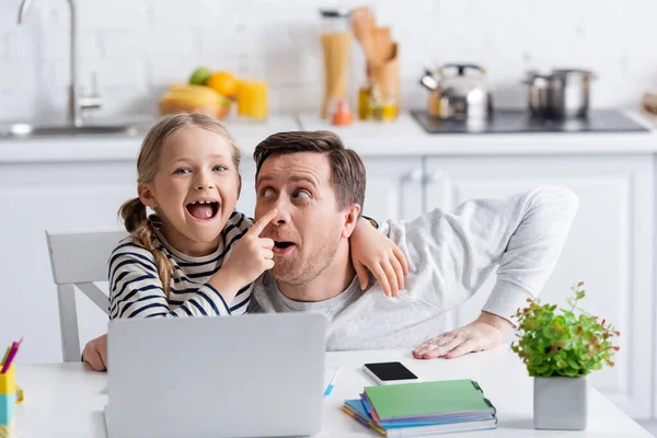 Excited girl touching nose of father while doing homework near laptop — Stock Photo