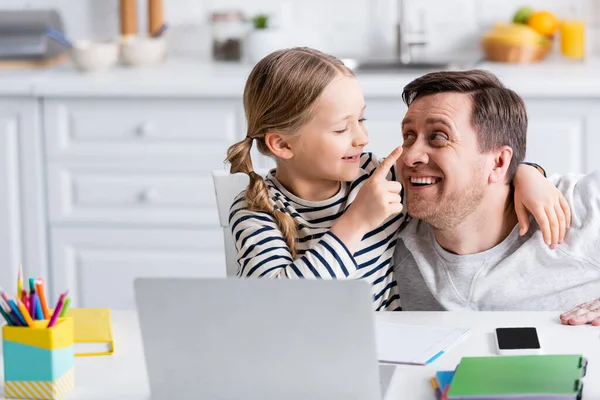 Alegre chica tocando la nariz de padre mientras se divierten cerca del ordenador portátil en la cocina - foto de stock