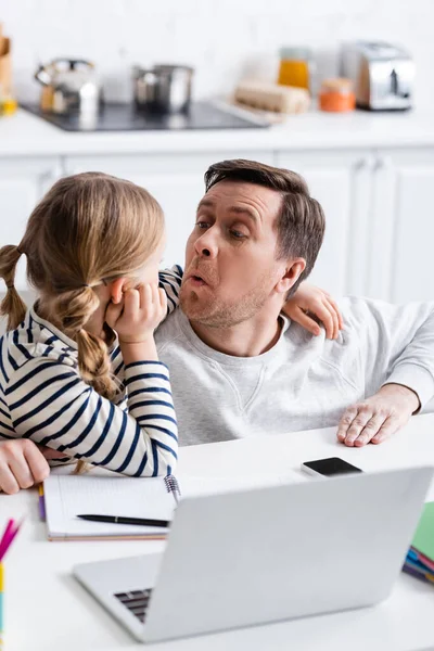 Padre haciendo muecas cerca de hija haciendo la tarea cerca de la computadora portátil en primer plano borrosa - foto de stock