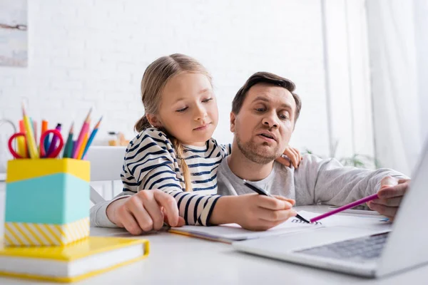 Vater und Tochter zeigen auf Notizbuch neben Laptop auf verschwommenem Vordergrund — Stockfoto