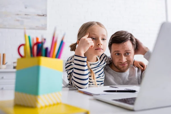 Padre reflexivo y la hija mirando a la computadora portátil durante la lección en línea en primer plano borrosa - foto de stock