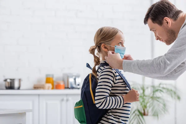 Side view of schoolgirl with backpack near father adjusting her medical mask — Stock Photo