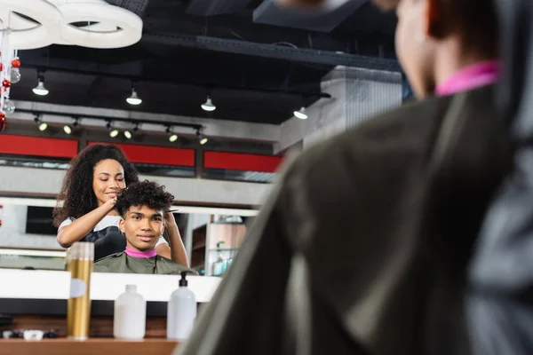 Lächelnder afrikanisch-amerikanischer Friseur steht neben Kundin im Umhang auf verschwommenem Vordergrund — Stockfoto