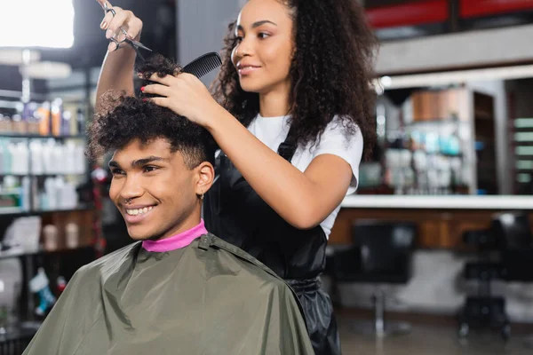 Alegre cliente afroamericano sentado cerca de peluquería sosteniendo tijeras y peine en el salón - foto de stock