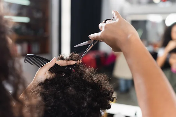 Vue recadrée de coiffeur afro-américain sur le premier plan flou tenant peigne et couper les cheveux de l'homme — Photo de stock