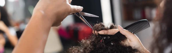 Обрезанный вид африканского американского парикмахера, держащего расческу и ножницы рядом с волосами клиента, баннер — стоковое фото