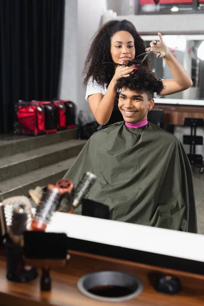 Parrucchiere afroamericano con forbici e pettine vicino ai capelli dell'uomo allegro — Foto stock