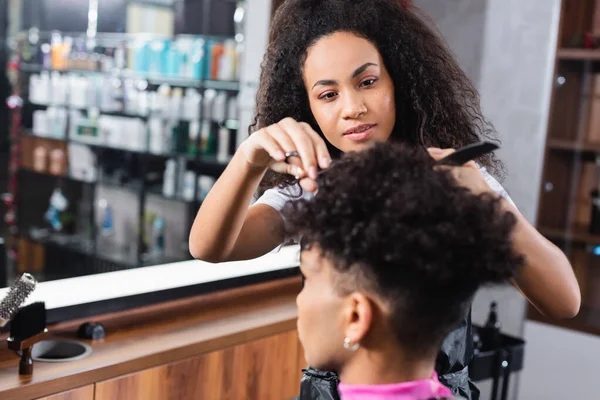 African american hairstylist cutting hair of man on blurred foreground in salon — Stock Photo