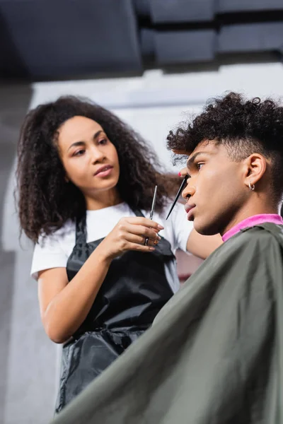 Vista de bajo ángulo del cliente afroamericano en capa sentada cerca de peluquería con tijeras sobre fondo borroso - foto de stock