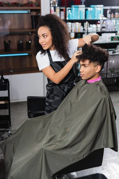 Smiling african american hairstylist combing hair of young man on chair in salon — Stock Photo