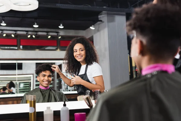 Smiling hairstylist holding comb near hair of african american client on blurred foreground — Stock Photo