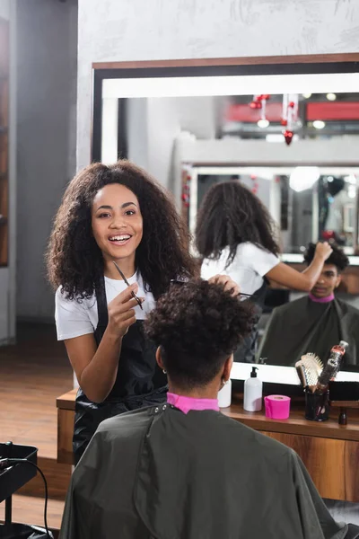 Peluquería afroamericana positiva con peine y tijeras trabajando con el cliente en el salón - foto de stock