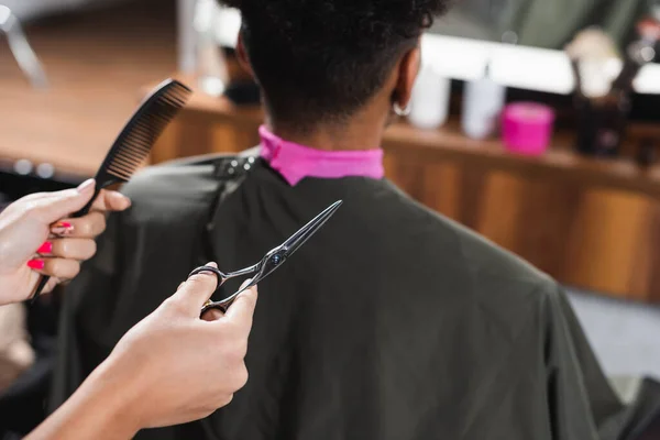 Scissors and comb in hands of hairdresser near african american man on blurred background — Stock Photo
