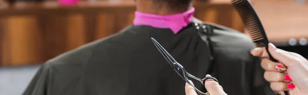Vista recortada de peluquería sosteniendo tijeras y peine cerca de cliente afroamericano, pancarta - foto de stock