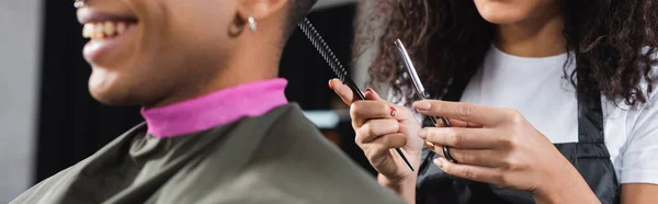 Vista recortada de la peluquería afroamericana sosteniendo tijeras y peine cerca de cliente sonriente en primer plano borroso, pancarta - foto de stock