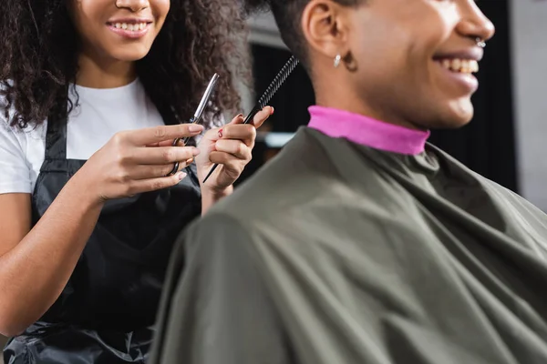 Vue recadrée du peigne et des ciseaux dans les mains du coiffeur afro-américain près d'un client souriant au premier plan flou — Photo de stock
