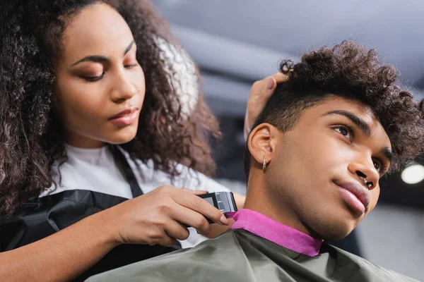 Low angle view of african american man sitting near hairdresser holding trimmer on blurred background — Stock Photo