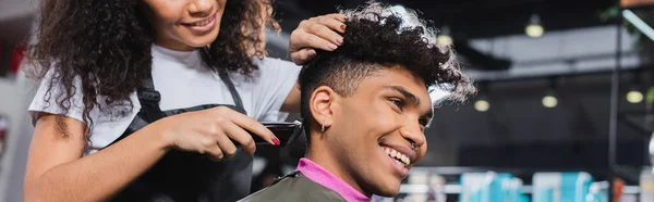 Sonriente hombre afroamericano sentado cerca de peluquería con trimmer en el salón, pancarta - foto de stock