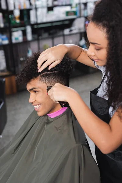 Cabeleireiro afro-americano em primeiro plano desfocado aparar o cabelo do homem sorridente — Fotografia de Stock