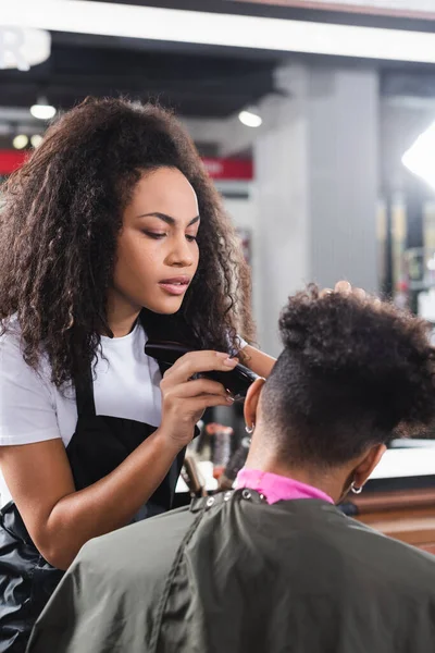 Focused african american hairstylist trimming hair of client in cape on blurred foreground — Stock Photo