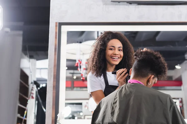 Africano americano cabeleireiro sorrindo para a câmera enquanto segurando aparador perto do homem no salão — Fotografia de Stock