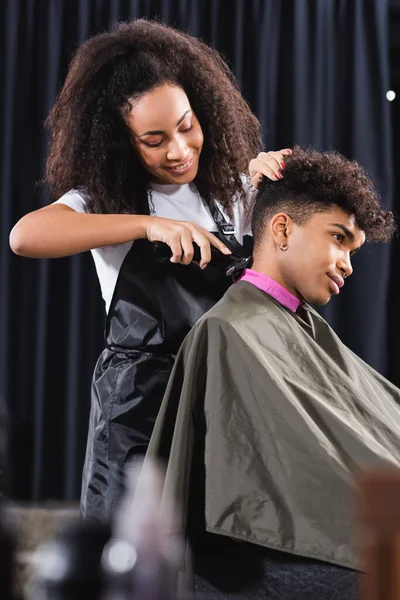 Happy african american hairdresser trimming neck of young man in cape and collar paper — Stock Photo