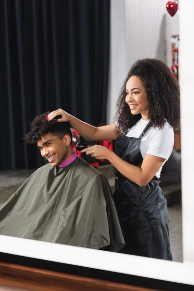 Alegre afroamericano hombre sentado cerca de peluquero en delantal celebración trimmer - foto de stock